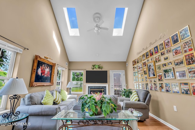 living area with a skylight, high vaulted ceiling, wood finished floors, baseboards, and ceiling fan