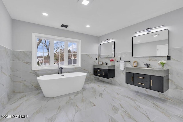 full bathroom with a sink, tile walls, two vanities, visible vents, and a soaking tub