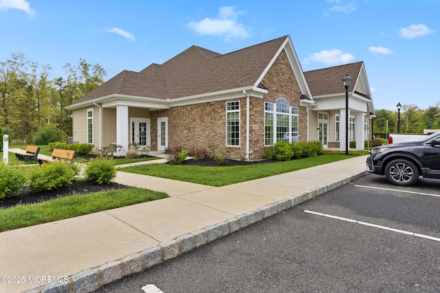 view of front facade featuring uncovered parking, stone siding, roof with shingles, a front lawn, and french doors