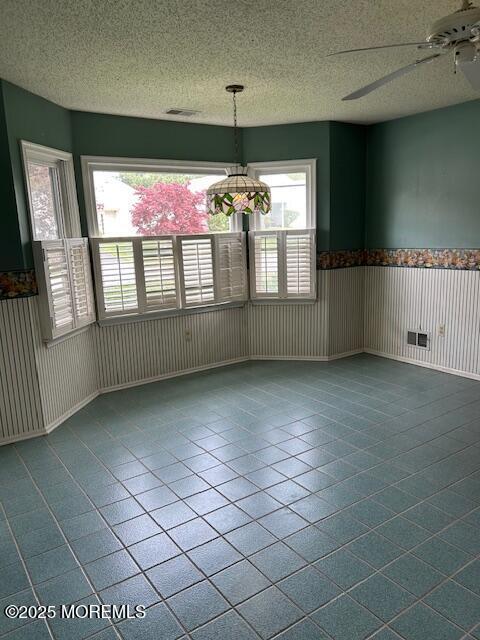unfurnished dining area featuring ceiling fan, wainscoting, a textured ceiling, dark tile patterned floors, and visible vents