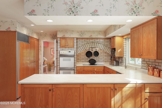kitchen with a peninsula, white double oven, a sink, wallpapered walls, and light countertops