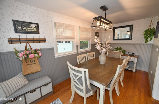 dining area featuring wallpapered walls, wood finished floors, a wainscoted wall, and a notable chandelier