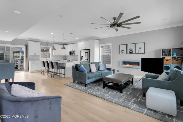 living area featuring ceiling fan, light wood-style flooring, crown molding, a glass covered fireplace, and recessed lighting