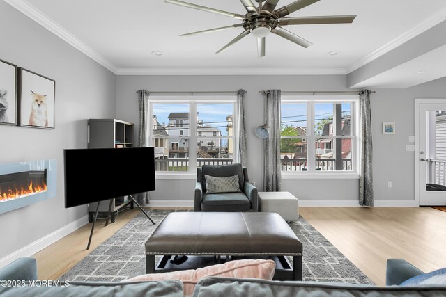 living area with ornamental molding, wood finished floors, ceiling fan, a glass covered fireplace, and baseboards