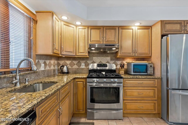 kitchen with appliances with stainless steel finishes, under cabinet range hood, a sink, light stone counters, and backsplash