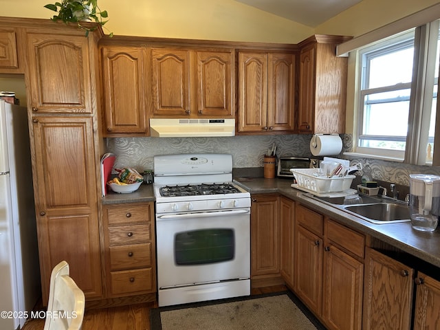 kitchen with white appliances, under cabinet range hood, brown cabinetry, a sink, and backsplash