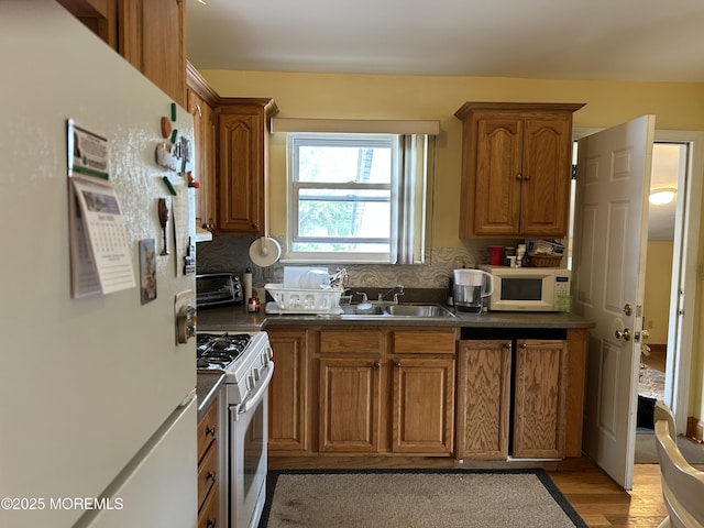 kitchen with white appliances, a sink, brown cabinetry, light wood-type flooring, and dark countertops
