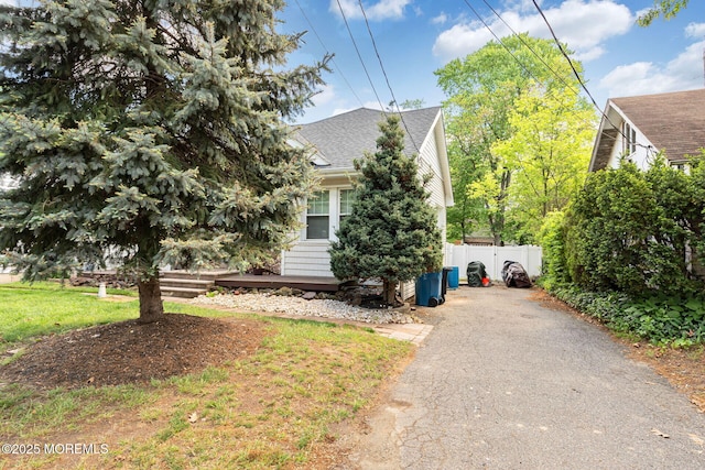 view of property hidden behind natural elements featuring a shingled roof, asphalt driveway, and fence