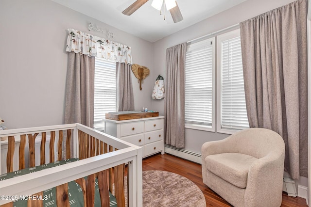 bedroom featuring baseboard heating, a nursery area, wood finished floors, and a ceiling fan