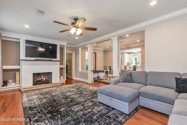 living area with ornate columns, ceiling fan, wood finished floors, ornamental molding, and a brick fireplace