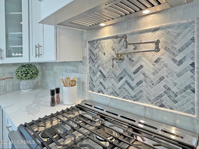 kitchen with wall chimney range hood, white cabinets, glass insert cabinets, cooktop, and light stone counters