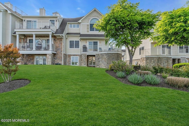 back of house featuring stone siding, a lawn, a balcony, and a chimney