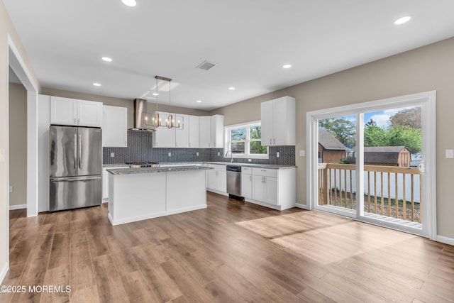 kitchen with appliances with stainless steel finishes, wall chimney range hood, backsplash, wood finished floors, and white cabinetry