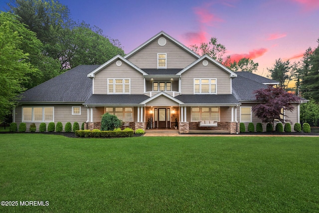 view of front of home featuring a lawn, roof with shingles, stone siding, and covered porch