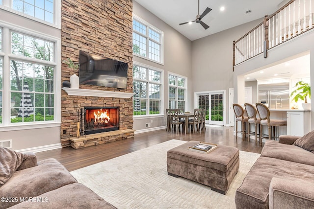 living area with a towering ceiling, ceiling fan, a stone fireplace, wood finished floors, and baseboards