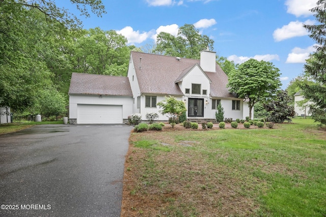 cape cod house featuring a chimney, a garage, asphalt driveway, roof with shingles, and a front lawn