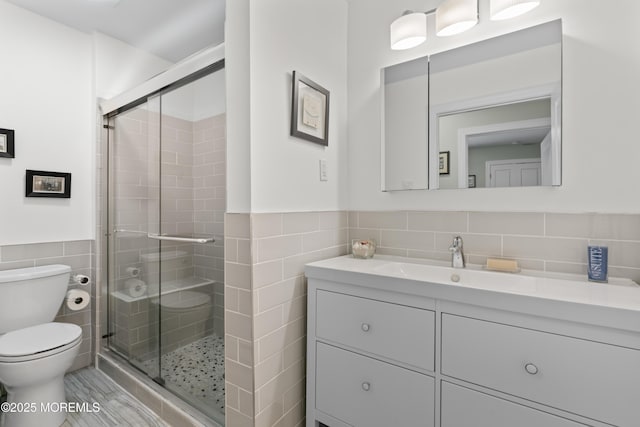 bathroom featuring tile walls, toilet, a stall shower, vanity, and a wainscoted wall