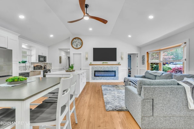 living area featuring a glass covered fireplace, lofted ceiling, a ceiling fan, light wood-style flooring, and a wainscoted wall