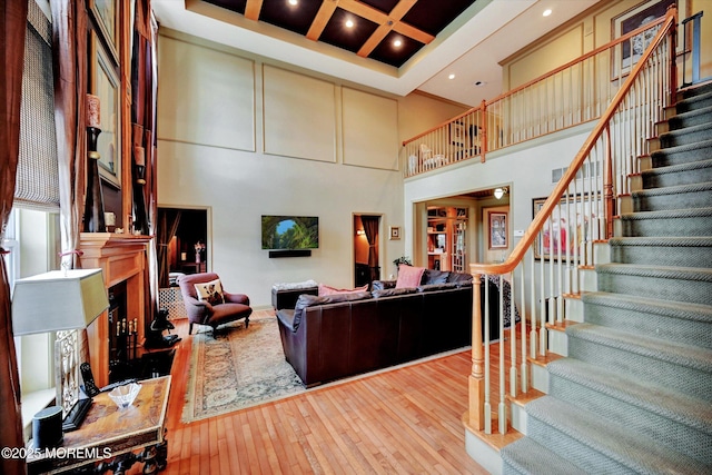 living area featuring a high ceiling, stairs, a fireplace with raised hearth, wood-type flooring, and coffered ceiling