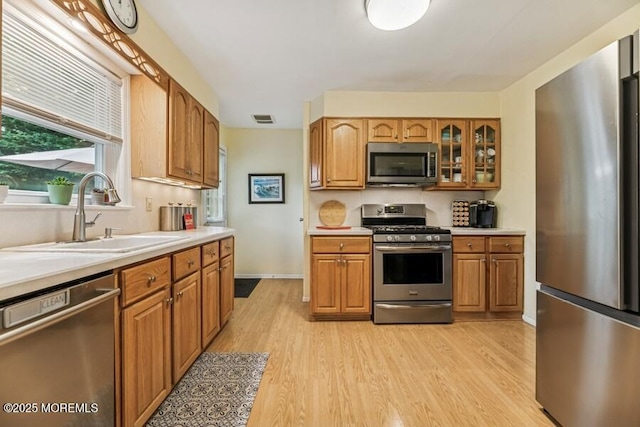 kitchen with appliances with stainless steel finishes, a sink, light countertops, light wood-style flooring, and glass insert cabinets