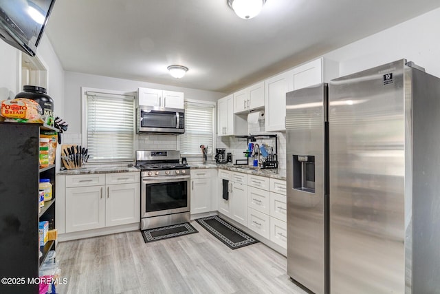 kitchen featuring appliances with stainless steel finishes, white cabinets, decorative backsplash, light stone counters, and light wood finished floors