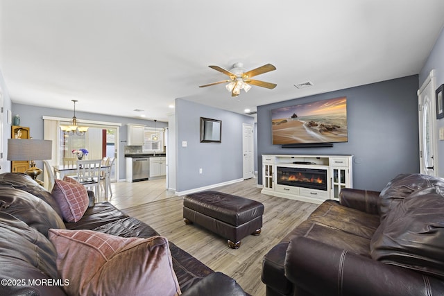 living room featuring a ceiling fan, light wood-style floors, a chandelier, baseboards, and a glass covered fireplace