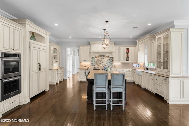 kitchen featuring double oven, cream cabinets, an island with sink, recessed lighting, and backsplash