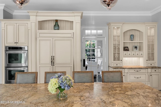 kitchen with double oven, cream cabinets, light stone counters, crown molding, and open shelves