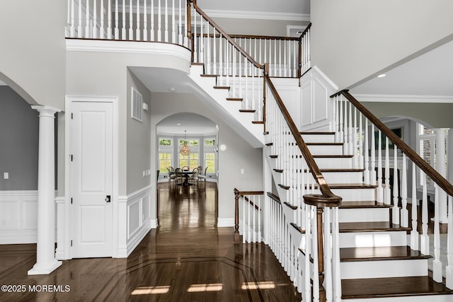 staircase with arched walkways, wood finished floors, ornamental molding, a high ceiling, and a wainscoted wall