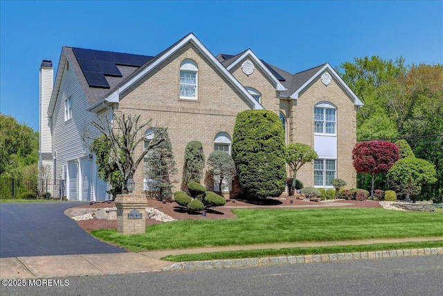traditional home with roof mounted solar panels, a front lawn, a chimney, brick siding, and driveway