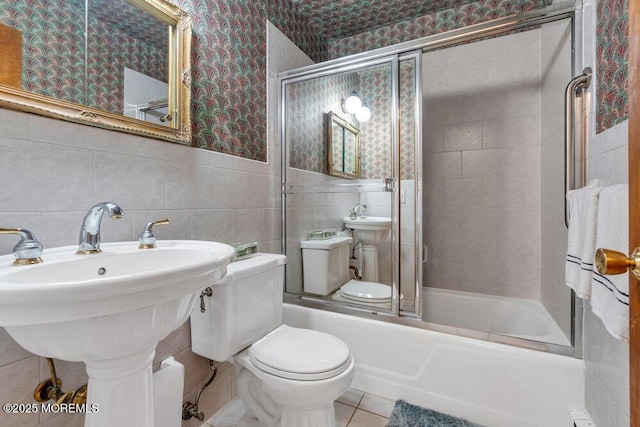 bathroom with wallpapered walls, toilet, bath / shower combo with glass door, tile patterned flooring, and tile walls