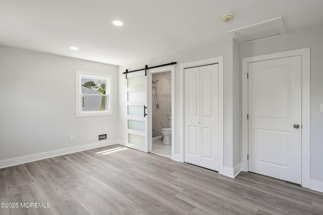 unfurnished bedroom with a barn door, baseboards, wood finished floors, ensuite bathroom, and attic access