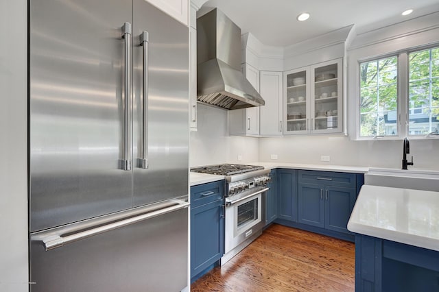 kitchen featuring high end appliances, blue cabinets, wall chimney exhaust hood, a sink, and glass insert cabinets