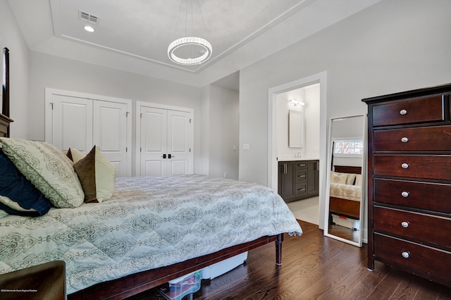 bedroom with a tray ceiling, two closets, dark wood-type flooring, a chandelier, and ensuite bath