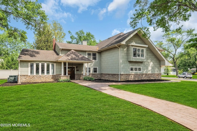 view of front facade with stone siding, a chimney, a front yard, a shingled roof, and central AC