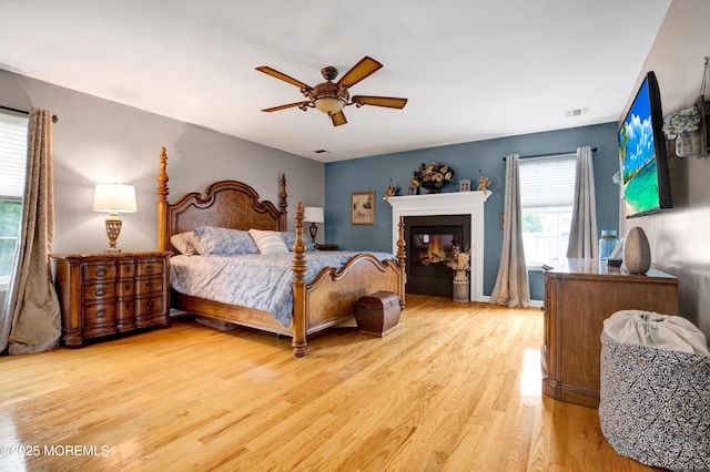 bedroom featuring light wood-style floors, a glass covered fireplace, and a ceiling fan