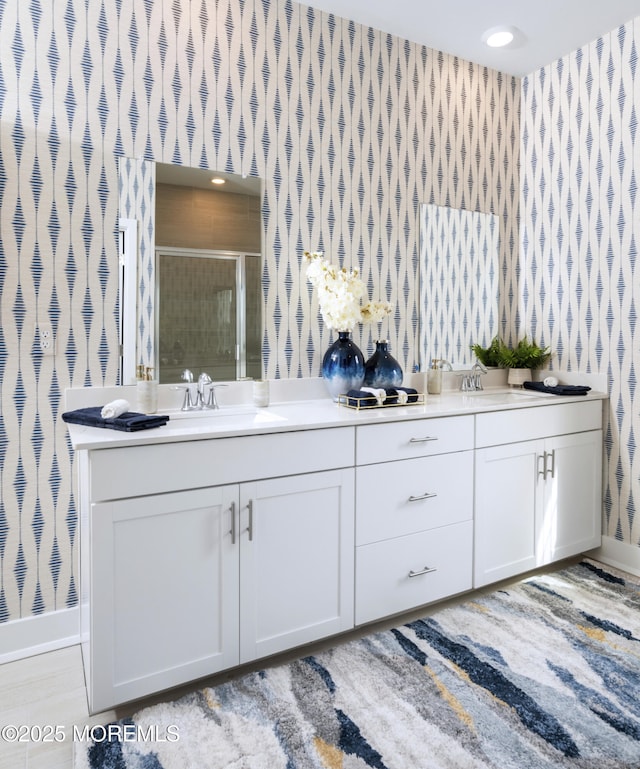 bathroom with wallpapered walls, baseboards, a shower stall, and double vanity