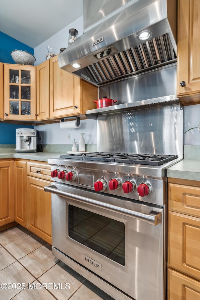 kitchen with designer range, wall chimney exhaust hood, light countertops, light tile patterned floors, and glass insert cabinets