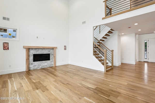 unfurnished living room featuring a high ceiling, light wood-type flooring, stairs, a stone fireplace, and baseboards