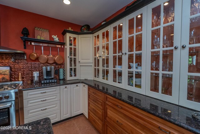 12 Hickory Dr, East Brunswick, NJ 08816 - kitchen featuring stainless steel range with gas stovetop, decorative backsplash, glass insert cabinets, light tile patterned floors, and dark stone counters