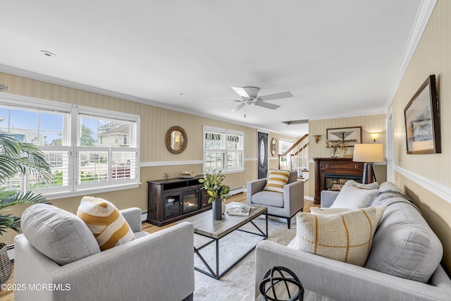 living area with a glass covered fireplace, crown molding, a baseboard heating unit, a ceiling fan, and light wood-type flooring