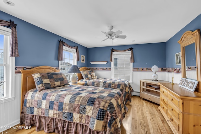 bedroom with light wood-type flooring, a wainscoted wall, a baseboard radiator, and a ceiling fan