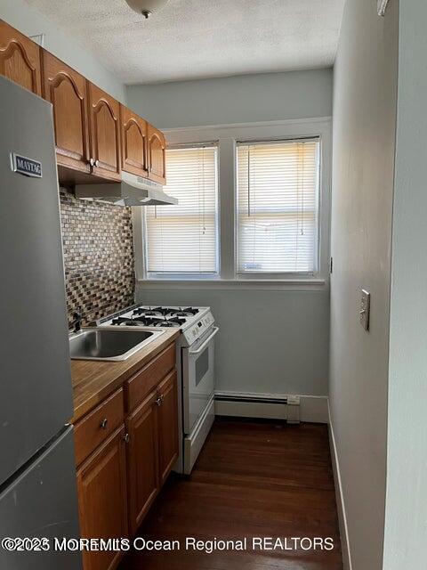 kitchen with baseboard heating, freestanding refrigerator, gas range gas stove, under cabinet range hood, and backsplash