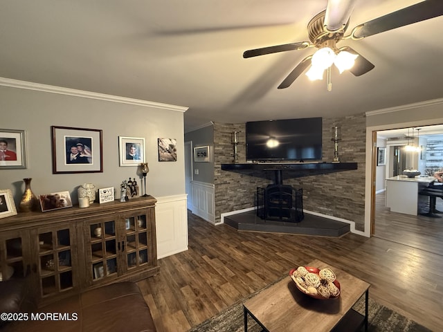 living room with ornamental molding, a ceiling fan, wood finished floors, a wood stove, and a wainscoted wall