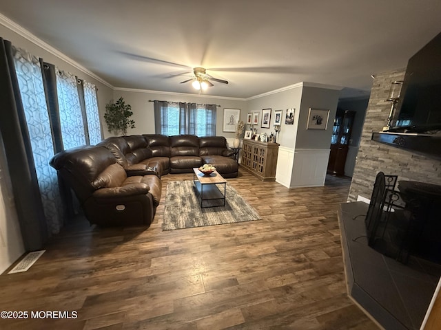 living area with a ceiling fan, crown molding, wood finished floors, wainscoting, and a stone fireplace
