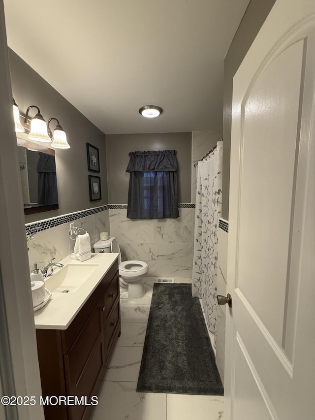 full bathroom featuring marble look tile flooring, toilet, vanity, tile walls, and wainscoting