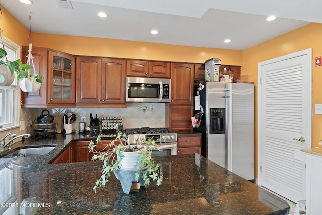 kitchen featuring appliances with stainless steel finishes, a sink, brown cabinetry, backsplash, and glass insert cabinets