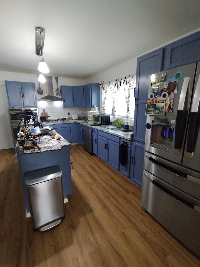 kitchen with wood finished floors, wall chimney range hood, appliances with stainless steel finishes, blue cabinetry, and tasteful backsplash