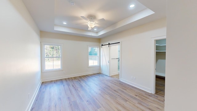 empty room with light wood-type flooring, a raised ceiling, ceiling fan, and a barn door