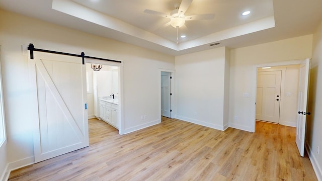 unfurnished bedroom with a raised ceiling, a barn door, ensuite bath, and light hardwood / wood-style flooring